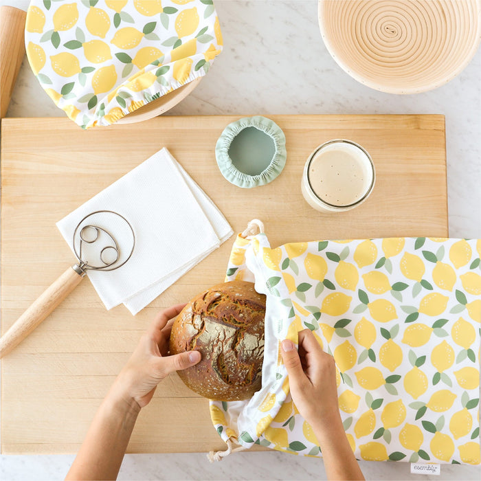 Person using a lemon-patterned reusable bread bag on a wooden table with bread and other items.