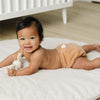 Baby lying on a white mattress with a toy, wearing a beige cloth diaper, in a room with a crib.