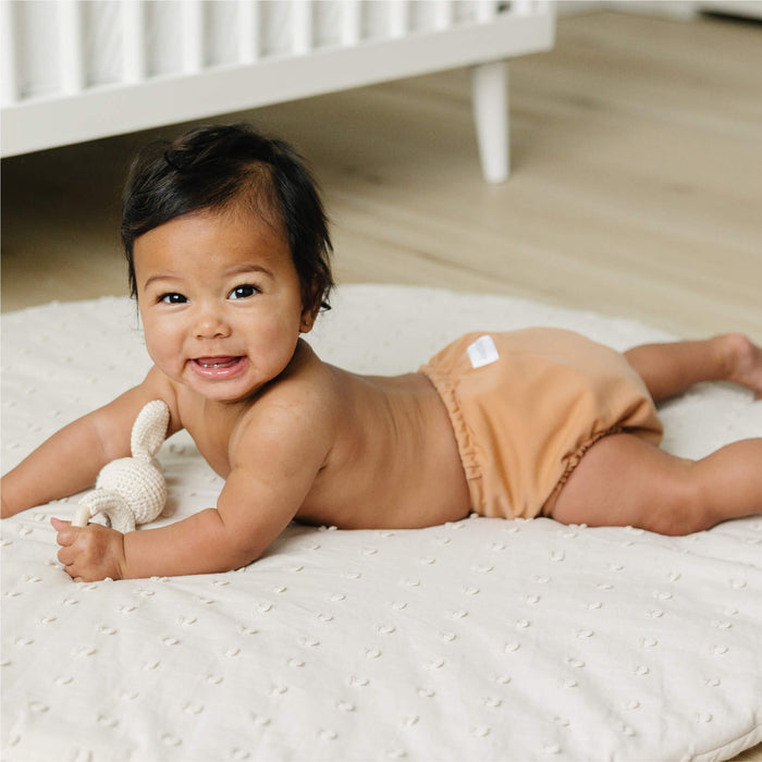 Baby lying on a white mattress with a toy, wearing a beige cloth diaper, in a room with a crib.