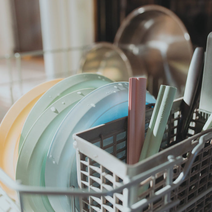 Two silicone straws (in pink and green) in a dishwasher next to silverware, plates, and bowls