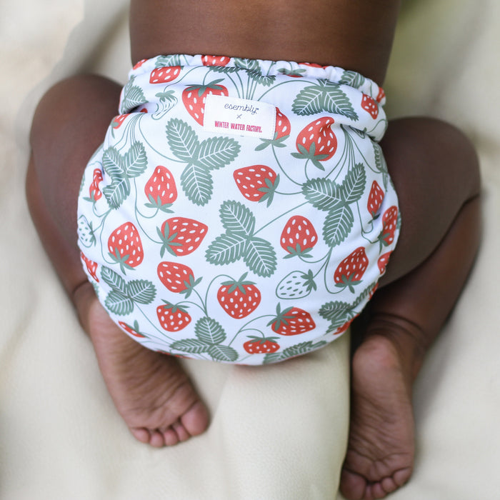 Baby laying on a white blanket wearing an Outer with a white background with red strawberries and light green leaves