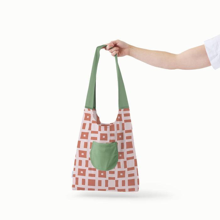 An outstretched arm holding an Esembly Day Bag in Theory (a reddish brown square and line geometric pattern on a light purple background with a mid-tone green strap and pocket)