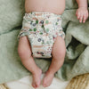 A baby wearing a patterned cloth diaper with a tractor-themed print, lying on a textured sage green blanket.