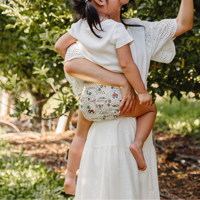 Mom holding a toddler wearing a Tractors Outer and picking apples in an orchard