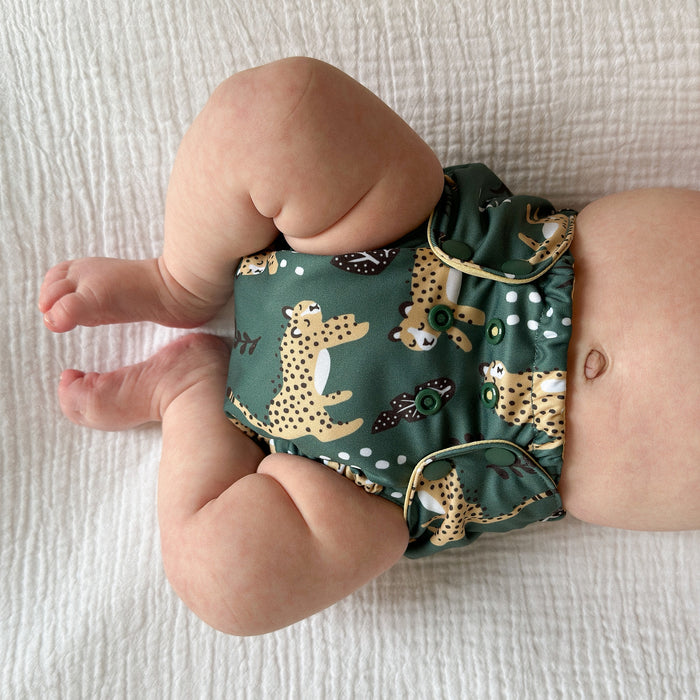Baby laying on a white blanket wearing Outer with a dark green background with yellow wildcat print and sporadic white dots