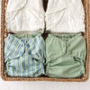 Two green cloth diapers with button closures and two stacks of Inners in a woven basket.