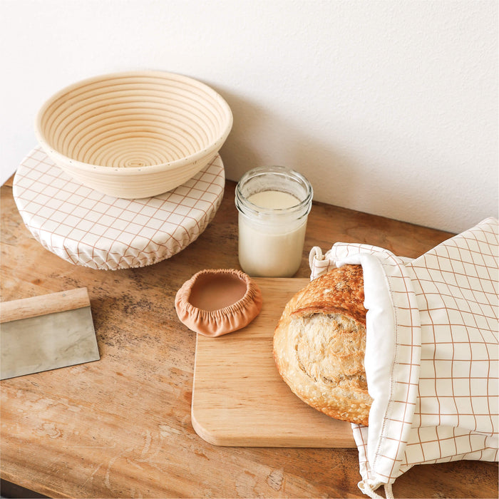 Bread in a Sourdough Set bag, jar of starter, and woven basket covered with a Bowl cap in Windowpane on a wooden table 
