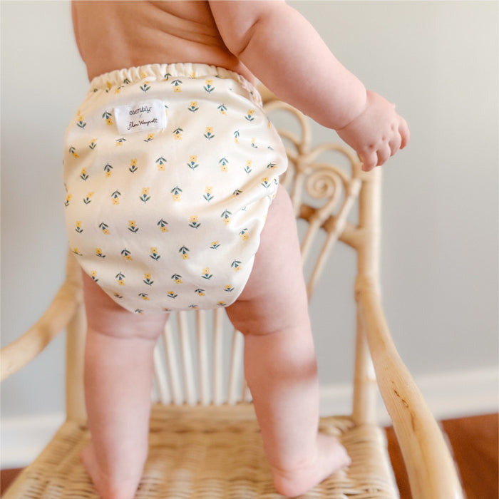 Baby standing on a wooden wicker chair wearing an Esembly Outer in Buds (a parchment background with tiny yellow and green flowers)