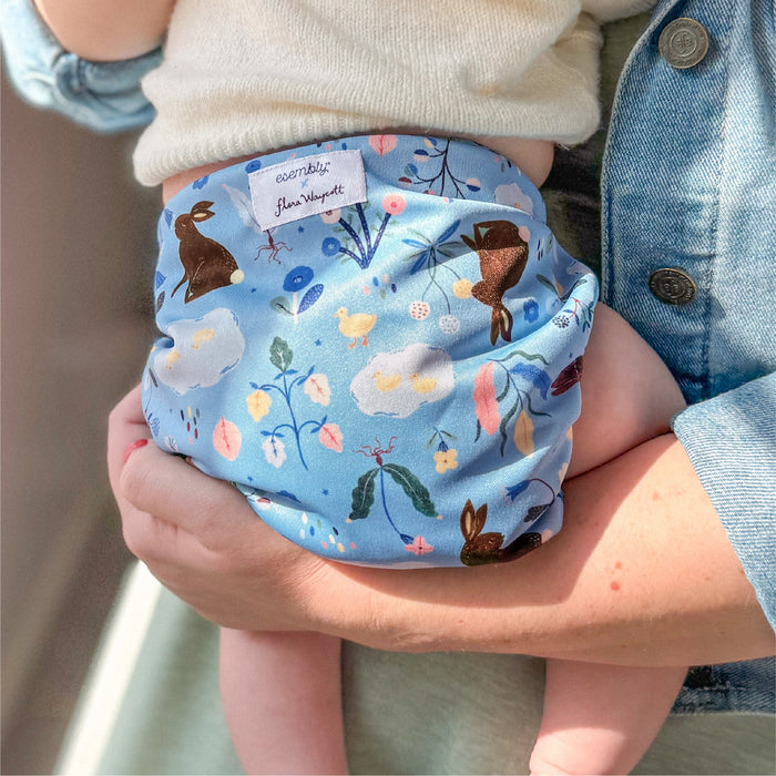A baby wearing a Bunnies (bunnies, flowers, and ducks illustrated by Flora Waycott on a mid-tone blue background) Esembly Outer being held by their mom