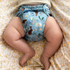 Baby laying on their back onto of a light pink and yellow floral sheet. The baby is wearing an Esembly Outer in Bunnies (bunnies, flowers, and ducks illustrated by Flora Waycott on a mid-tone blue background)