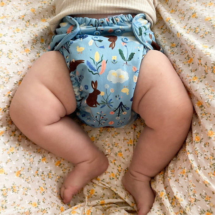 Baby laying on their back onto of a light pink and yellow floral sheet. The baby is wearing an Esembly Outer in Bunnies (bunnies, flowers, and ducks illustrated by Flora Waycott on a mid-tone blue background)