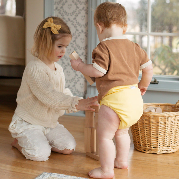 Siblings playing with wooden blocks. One of them is wearing an Esembly Outer in Buttercup (a buttery mid-tone yellow).