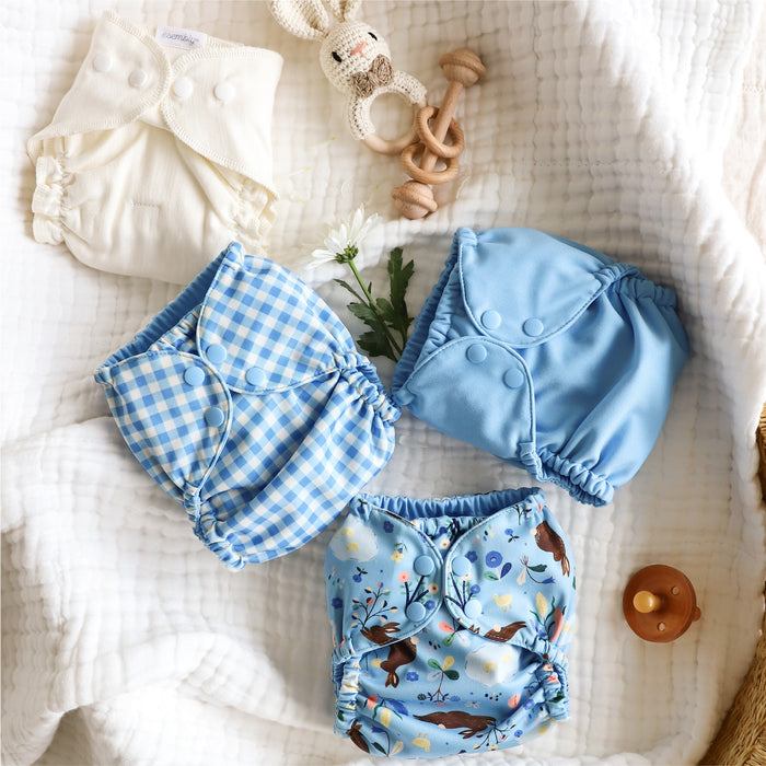 An Esembly Inner and three Outers (in blue Gingham, Cornflower (a soft, mid-tone blue), and Bunnies (bunnies, flowers, and ducks illustrated by Flora Waycott) on top of a white gauze blanket in a basket.