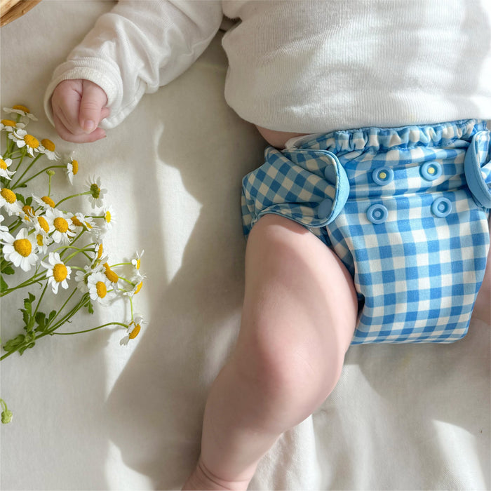 Baby laying on their back next to flowers wearing a blue Gingham Esembly Outer